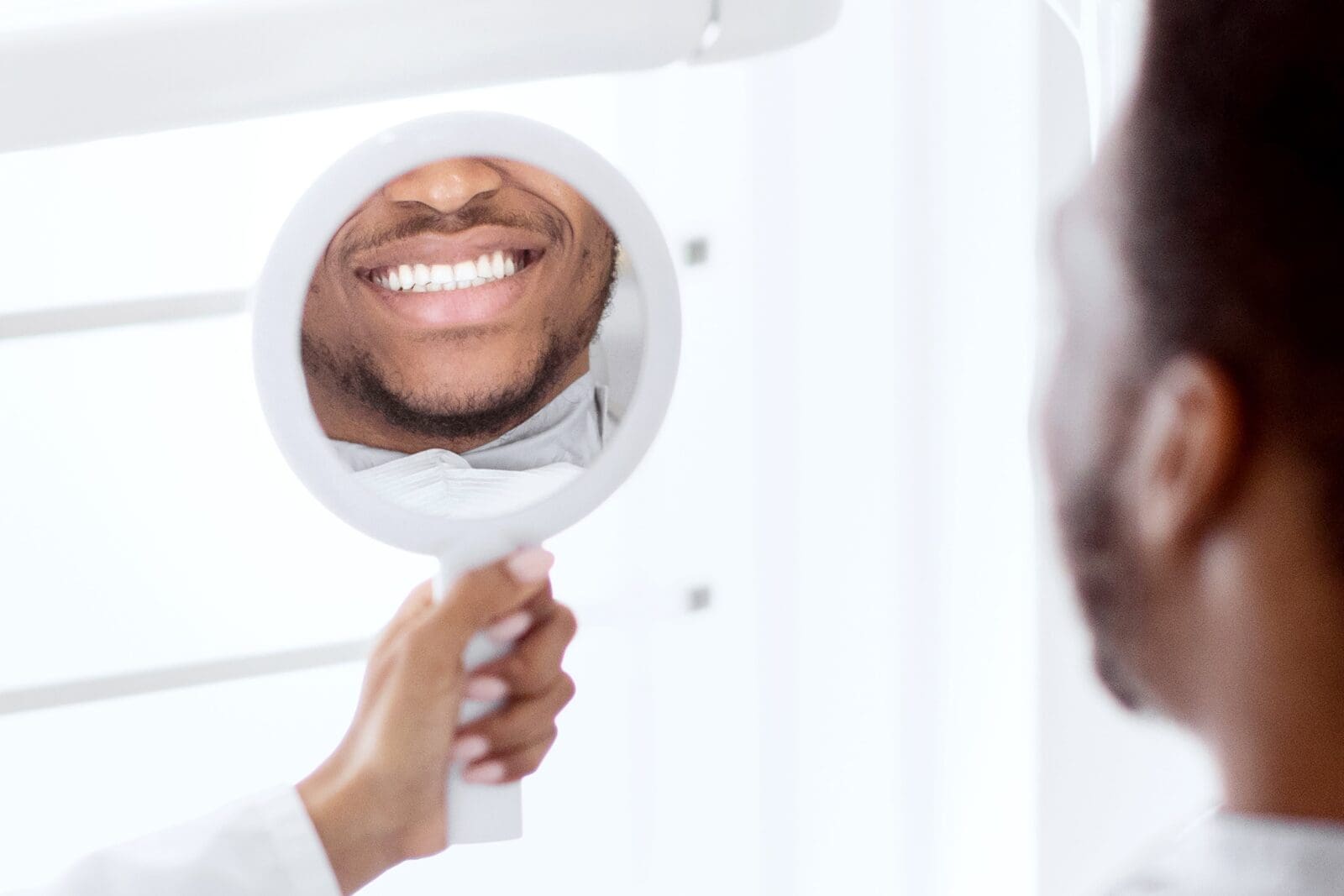 Black Guy Looking At Mirror, Checking His Teeth After Dental Treatment
